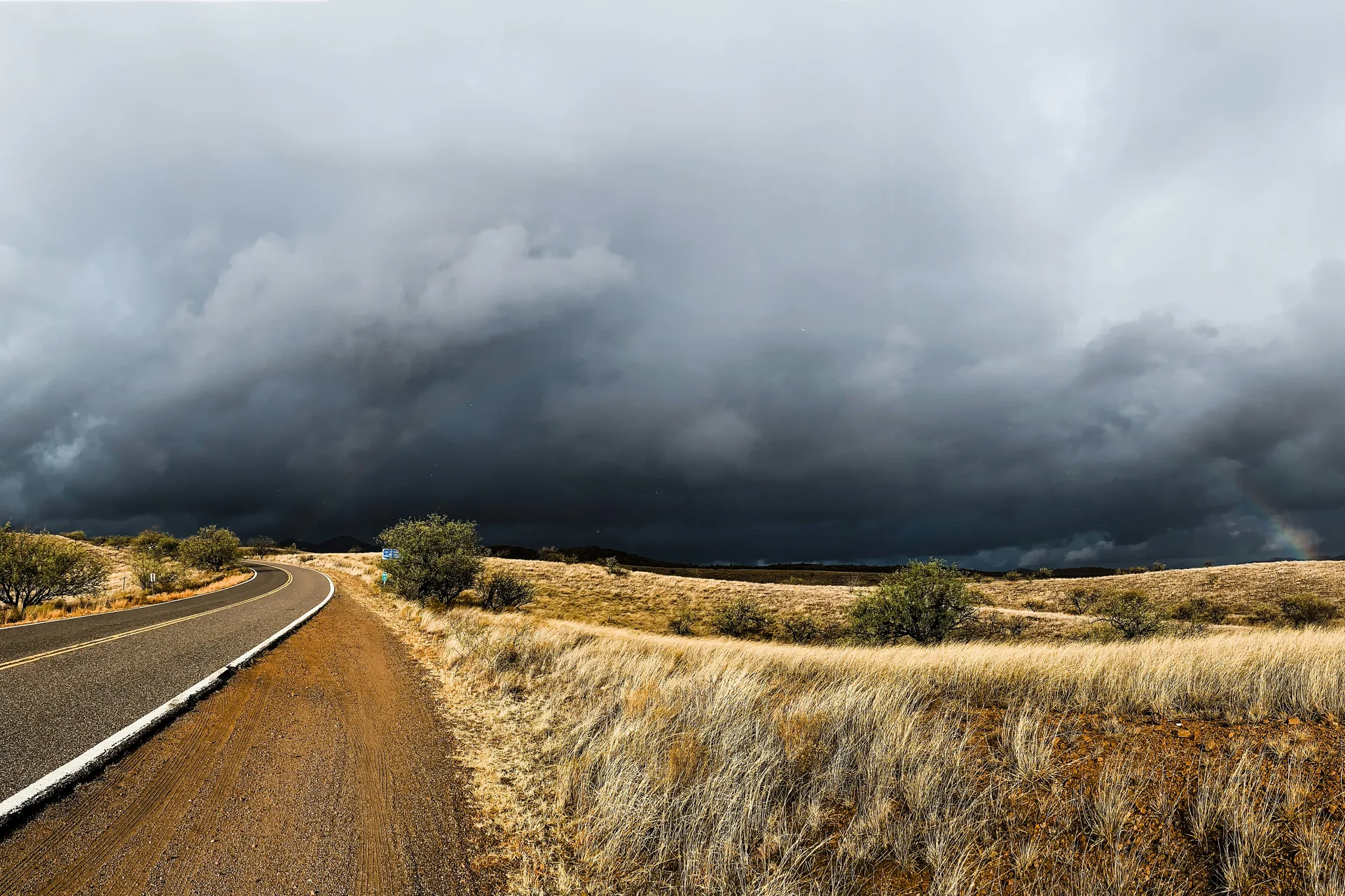 Dark storm clouds over a winding road and dry grassland, with a faint rainbow on the horizon